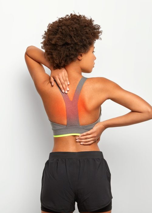Spine pain or backache concept. Horizontal shot of slim dark skinned woman has cronic painful feelings in back, poses against white background, has injury or cramps, wears grey top and shorts